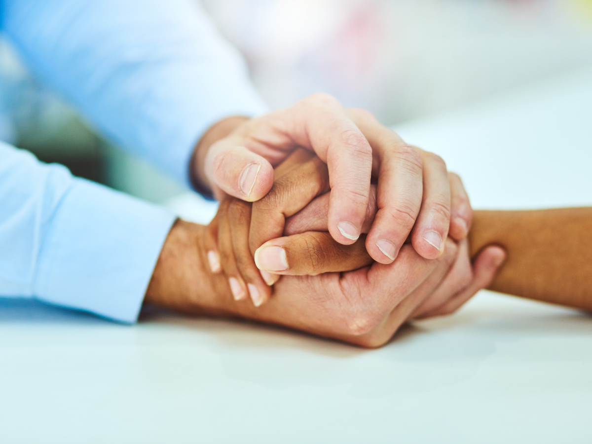 Two hands gently hold one another over a white surface. One hand is positioned on top in support, while the other is underneath. The background is softly blurred.