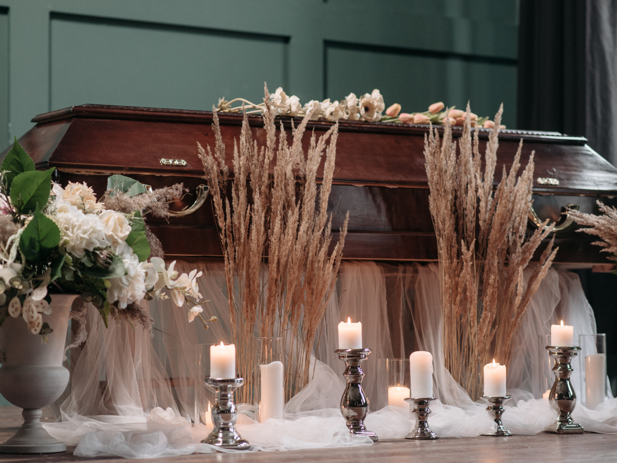 A wooden casket decorated with flowers, surrounded by white candles, dried grasses, and white fabric, sits in a solemn room with dark green paneled walls.
