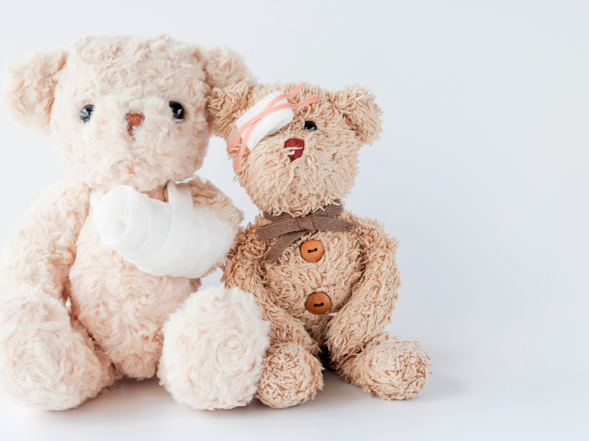 Two teddy bears are sitting side by side. One has a bandaged arm in a sling, and the other has a bandage wrapped around its head and over one eye. Both have a soft, well-loved appearance against a plain white background.