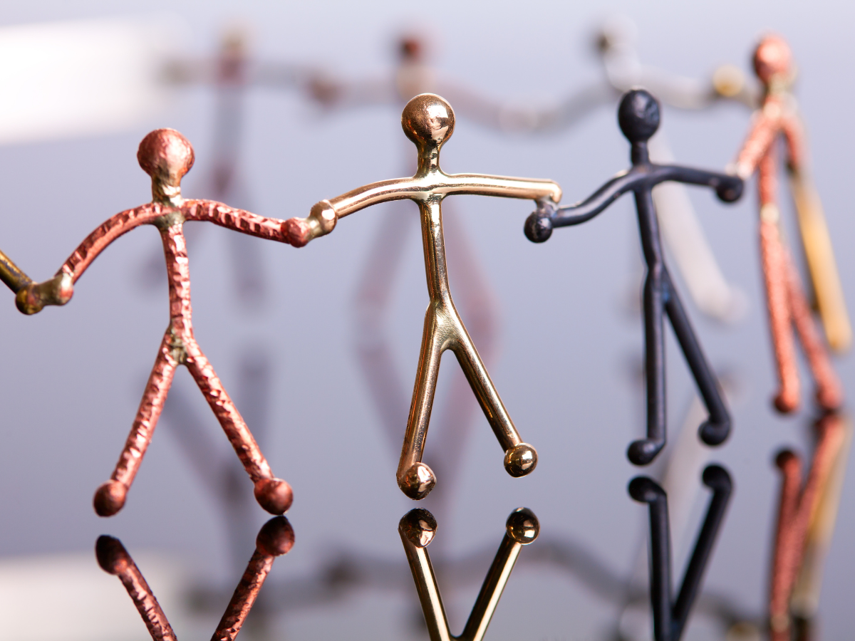 Five metallic stick-figure sculptures in different colors hold hands, forming a chain. Their reflections are visible on the shiny surface below them. The background is softly blurred.