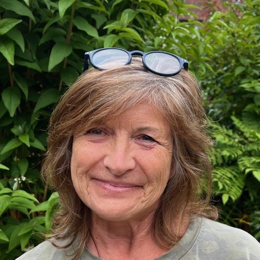Karen Woodger, a smiling older woman with shoulder-length brown hair, stands outdoors in front of green foliage, wearing glasses on top of her head and a green shirt.