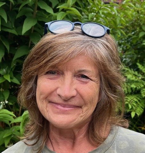 Karen Woodger, a smiling older woman with shoulder-length brown hair, stands outdoors in front of green foliage, wearing glasses on top of her head and a green shirt.