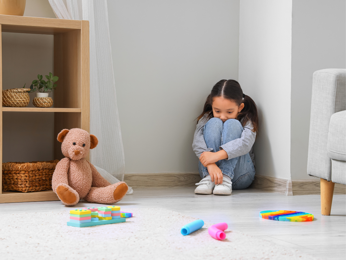 A young girl sits alone in the corner of a room, hugging her knees and looking sad. Nearby are a teddy bear, baskets on a shelf, and scattered colorful toys on a white carpet.