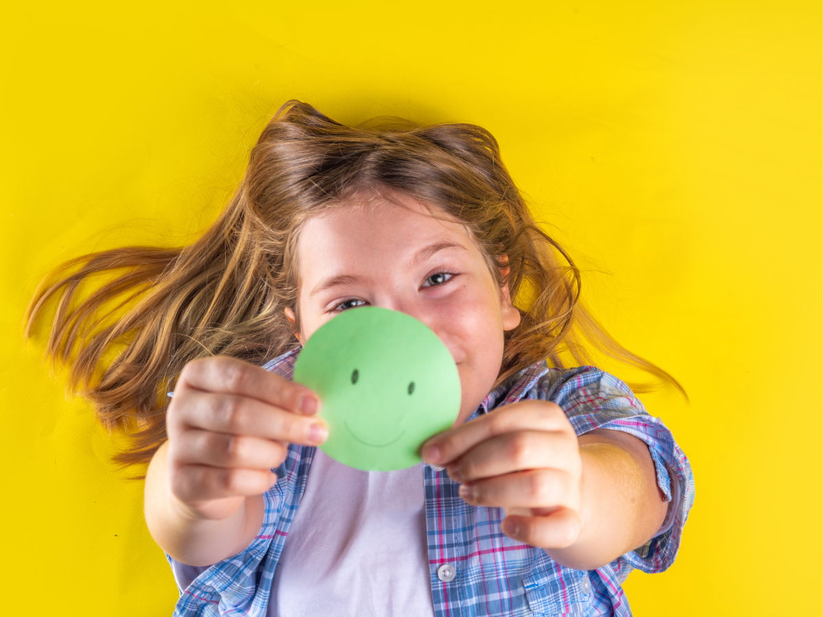 A child lies on a yellow background, smiling and holding a green paper circle with a smiley face drawn on it toward the camera. The child has long hair and is wearing a plaid shirt over a white t-shirt.