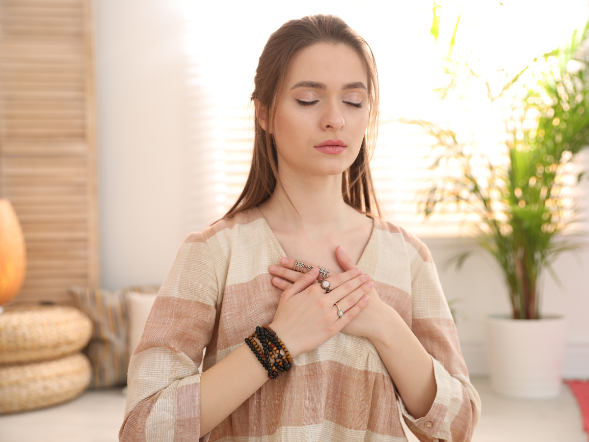 A woman with closed eyes stands indoors, holding her hands over her chest, wearing bracelets and a striped beige top, appearing calm and meditative, with soft sunlight and a potted plant in the background.
