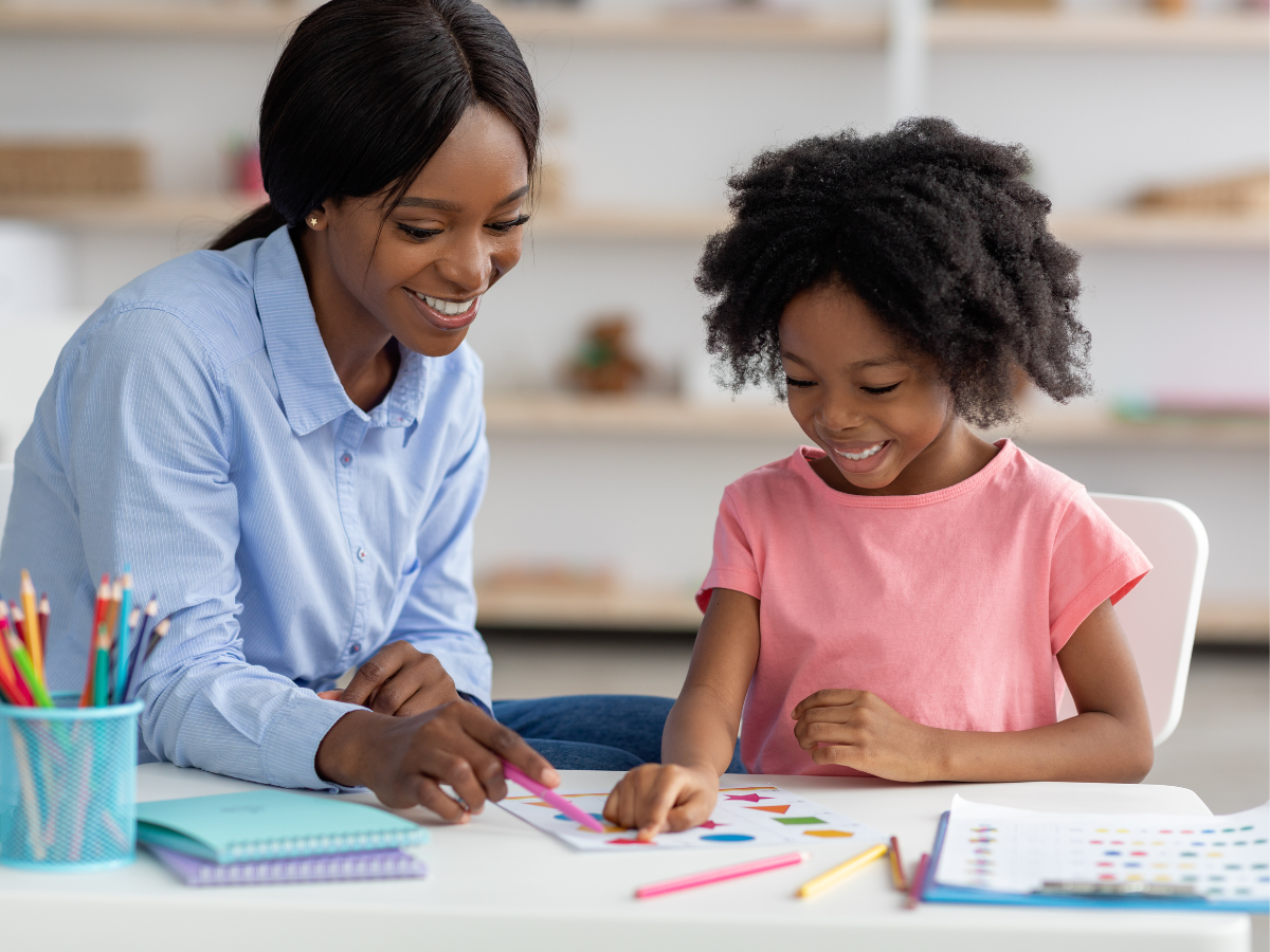 A woman and a young girl sit at a table, smiling as they draw with colored pencils. Art supplies and notebooks are on the table, and the setting looks bright and cheerful.