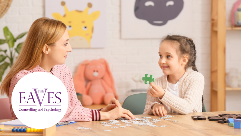 A woman and a young girl sit at a table working on a jigsaw puzzle. The girl holds up a puzzle piece and smiles. The background shows stuffed animals, and the Eaves Counselling and Psychology logo is in the foreground.
