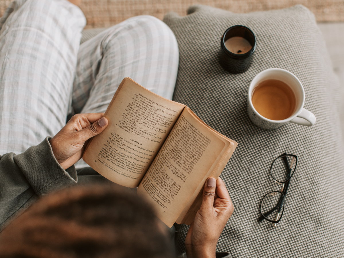 Person reading a book on a sofa with tea and glasses nearby, enjoying a calm moment for stress relief.