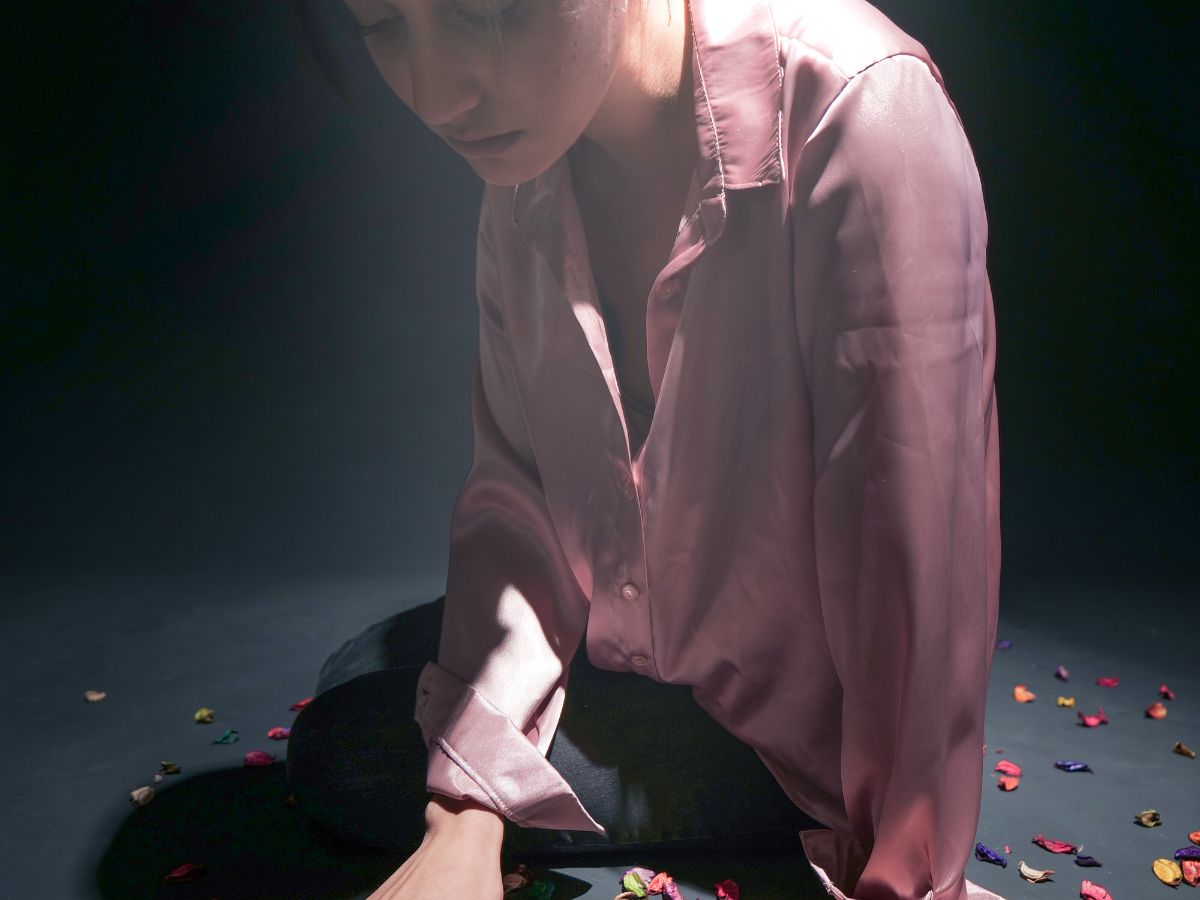 Woman in a pink satin blouse sitting on a dark floor, illuminated by a single overhead light, looking down in despair, representing emotional struggle and trauma.