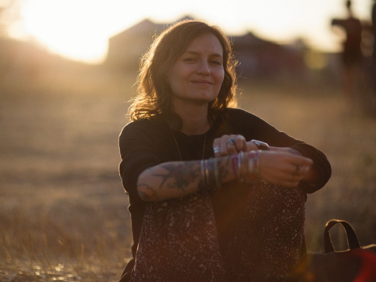 Woman sitting with legs pulled up, arms around them, smiling softly in soft sunset light with an open landscape, representing peace, hope, and trauma recovery.