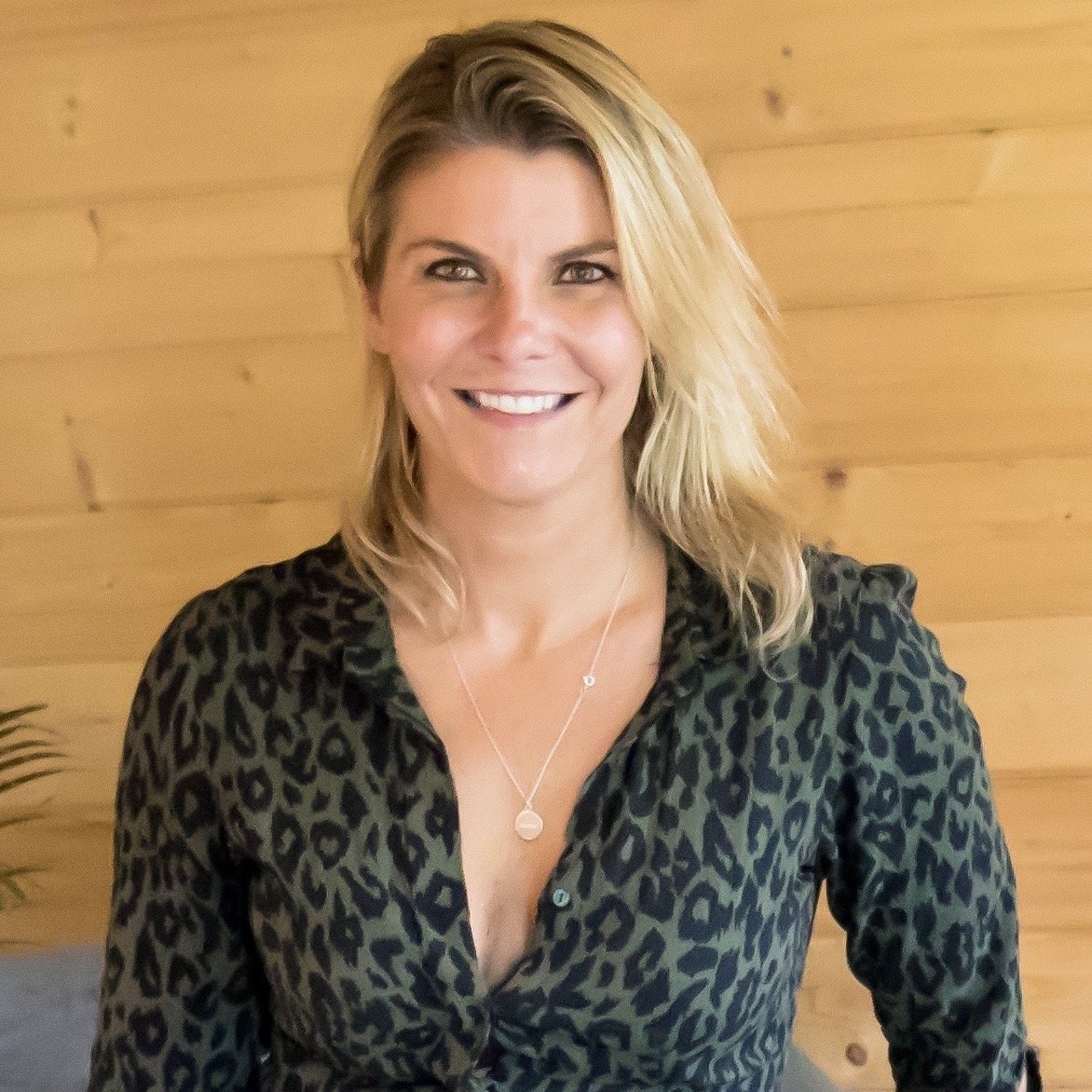 A woman with blonde hair wearing a green and black leopard print blouse and a necklace, smiling in front of a light wooden wall.