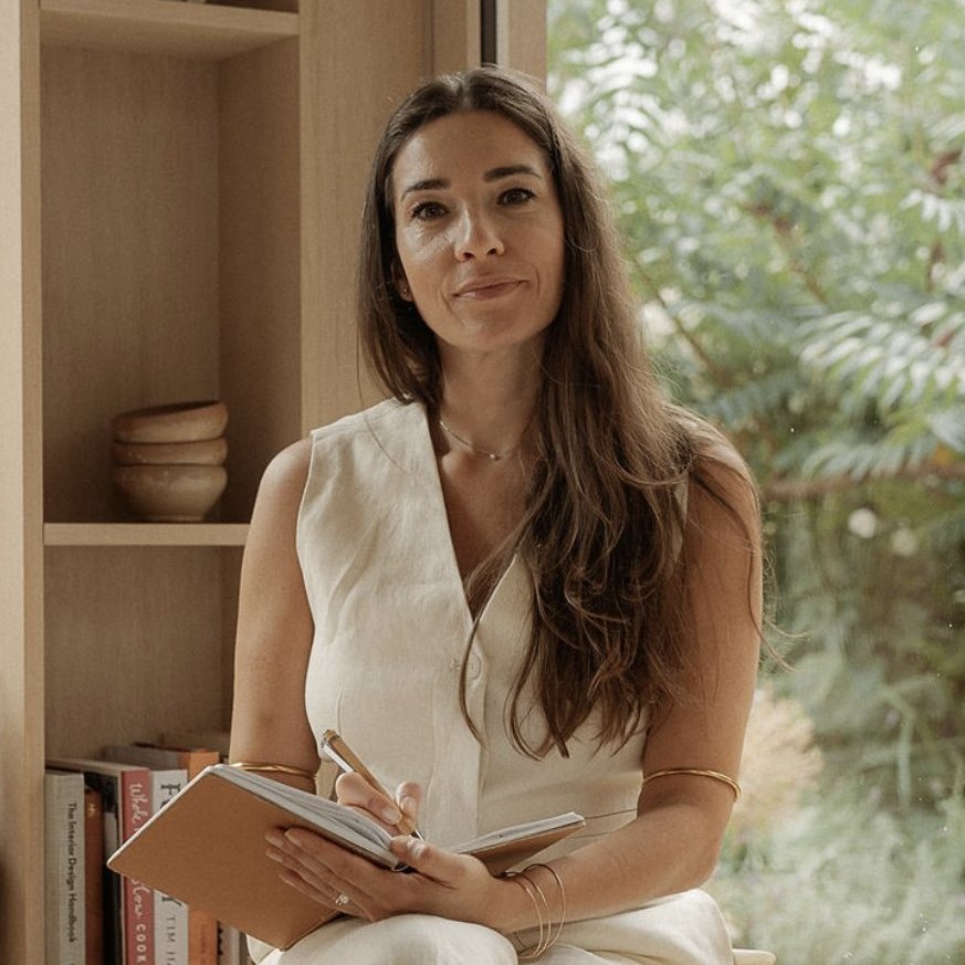 A woman with long brown hair, wearing a sleeveless beige outfit, sits by a window holding a notebook and pen, smiling softly. Behind her are wooden shelves with books, bowls, and a decorative sculpture. Greenery is visible outside.