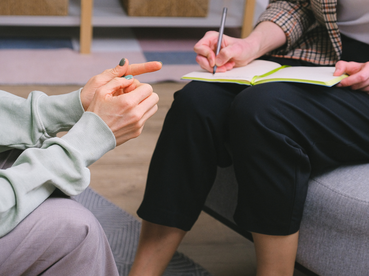 Two people sitting together; one is gesturing with their hands while the other takes notes in a notebook, suggesting a conversation or counseling session.