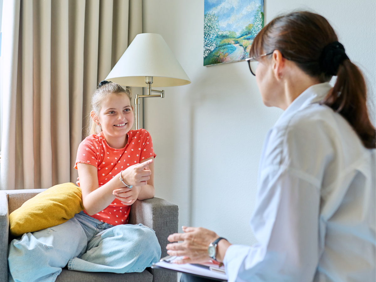 A smiling young girl in a red polka dot shirt sits on a couch talking to a woman in a white coat, who is holding a clipboard. They appear to be having a friendly conversation in a bright room.