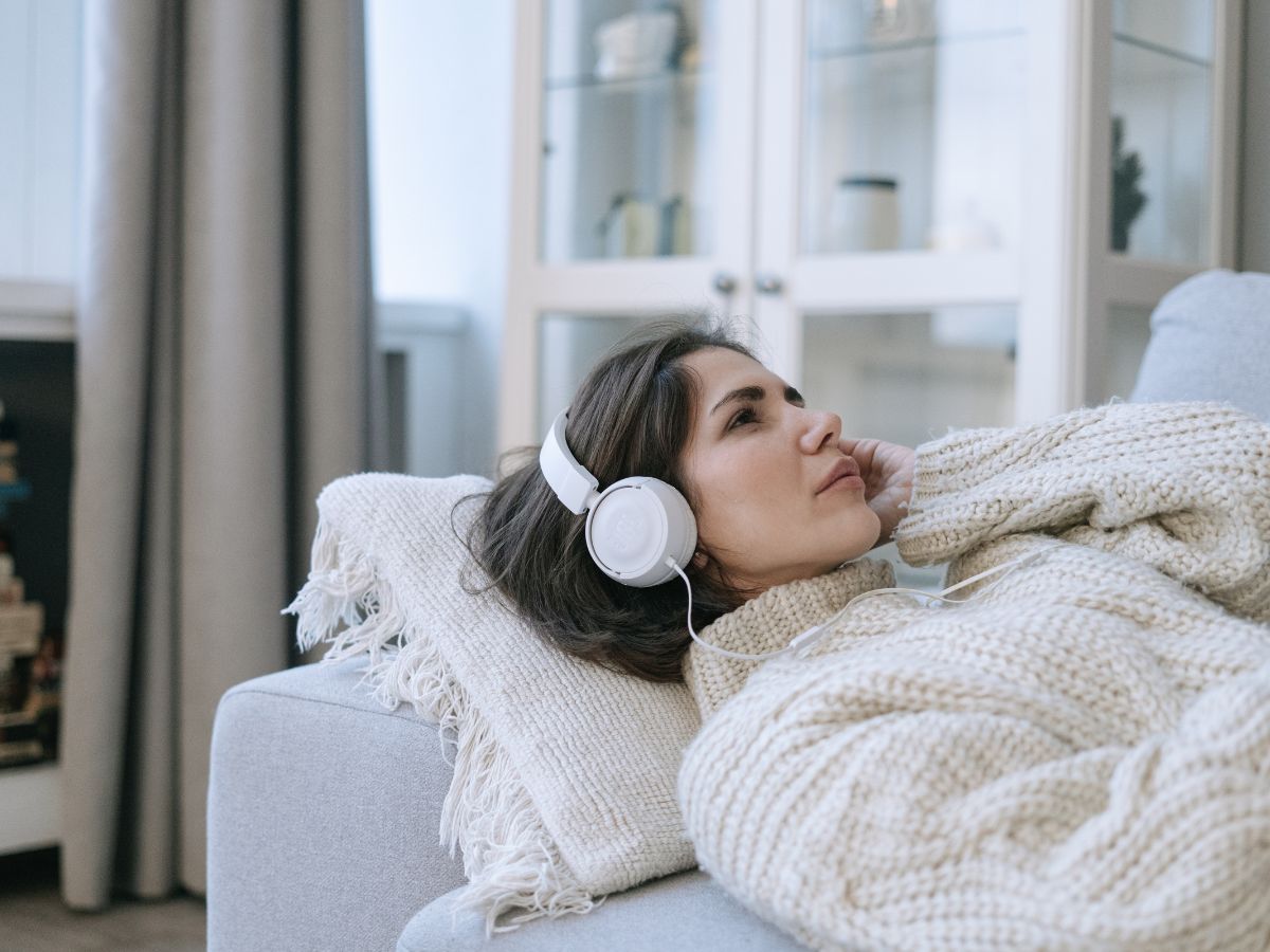 Woman lying on a sofa wearing headphones in a calm, neutral white living space, representing rest, emotional processing, and self-care during empty nest syndrome and grief.