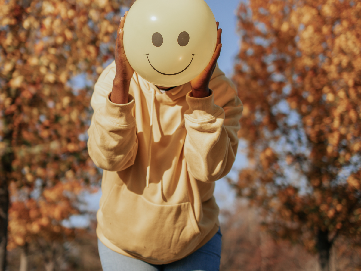 Young girl outdoors in autumn holding a yellow smiley-face balloon in front of her face, wearing jeans and a yellow hoodie