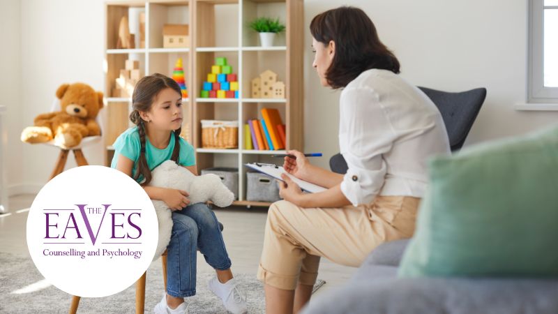 Child psychologist talking with a young girl holding a teddy bear in a child-friendly therapy room with toys and books