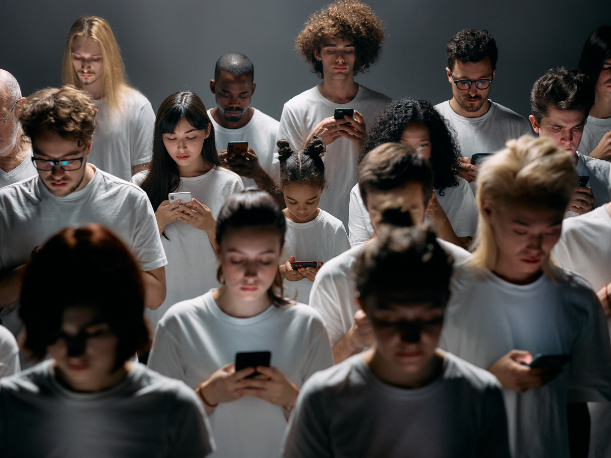 Dark room with a large group of people standing under dim overhead light, all looking at their smartphones like zombies