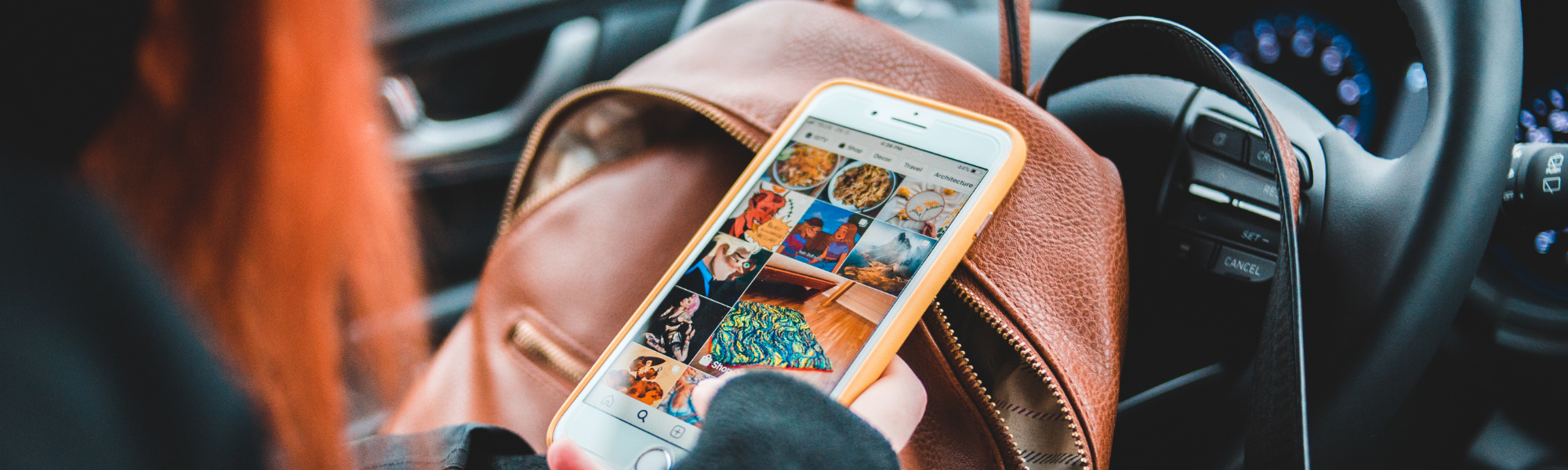 Woman sitting in a car looking at her smartphone, brown leather backpack on her lap, viewed from behind with steering wheel in background