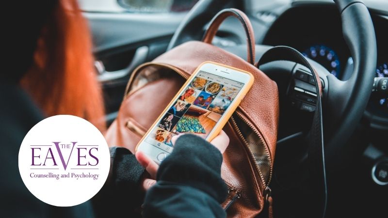 Woman sitting in a car looking at her smartphone, brown leather backpack on her lap, viewed from behind with steering wheel in background