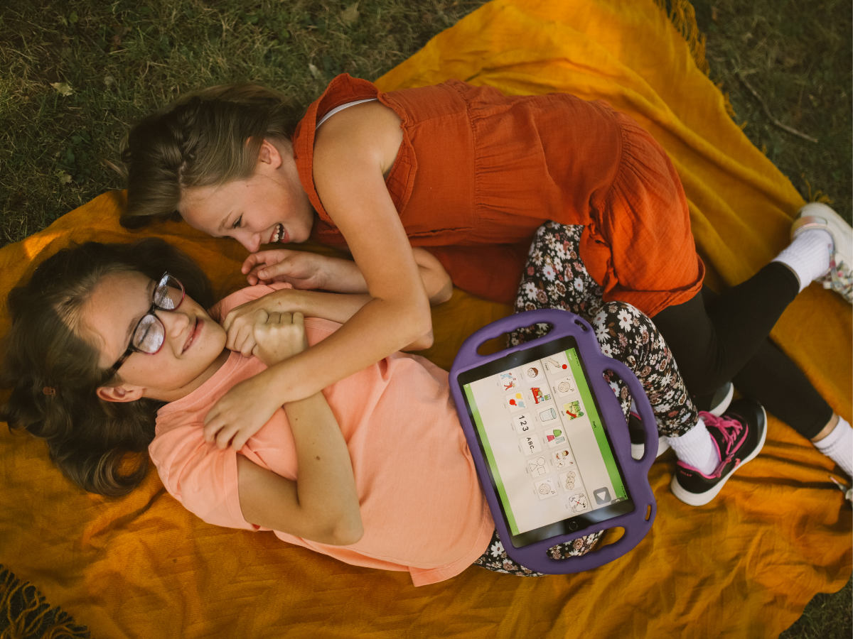 Two young girls lying on a blanket outdoors, engaging in playful interaction while using a tablet, supporting social connection and emotional development