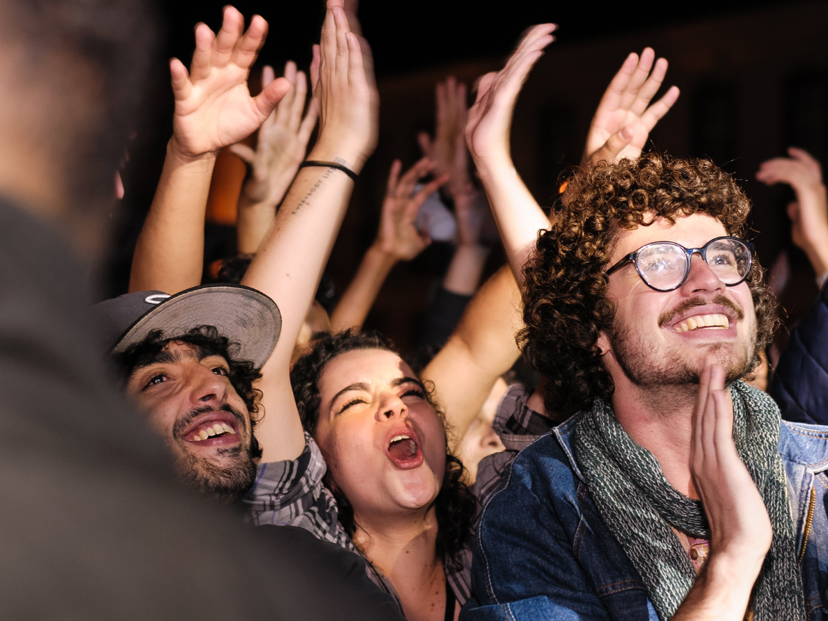 Three concertgoers cheering and clapping hands in the foreground, with more hands raised from the crowd behind them at a live music event.
