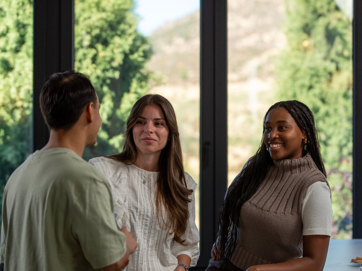 Three friends – one man and two women – smiling and having a casual indoor conversation near glass doors with a forest outside, symbolizing connection, joy, and healthy friendships.