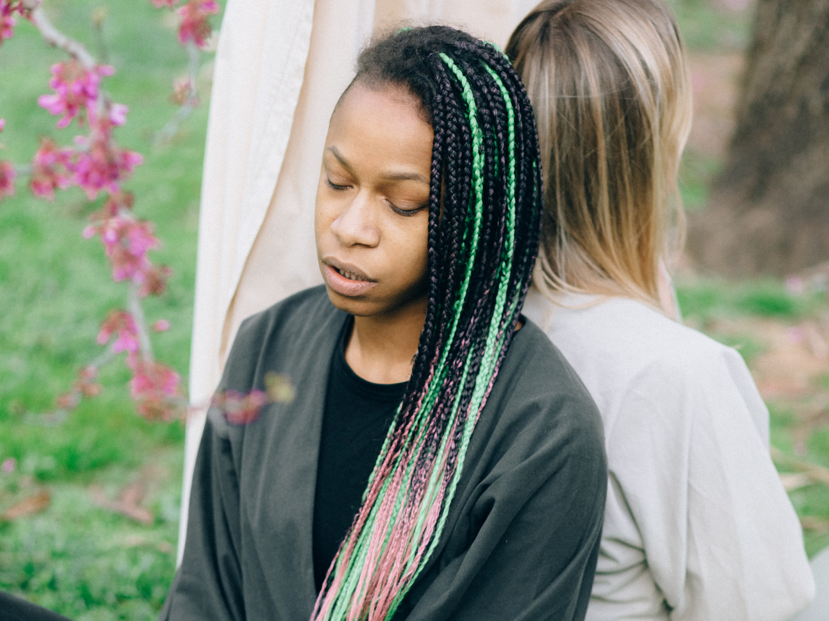 Two women sitting back-to-back on grass, looking sad or withdrawn, representing emotional struggle, social overwhelm, or feeling disconnected.