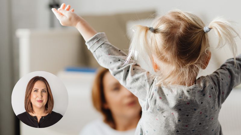 Toddler raising hands while mom sits on the floor in the background, illustrating parental burnout and parenting challenges