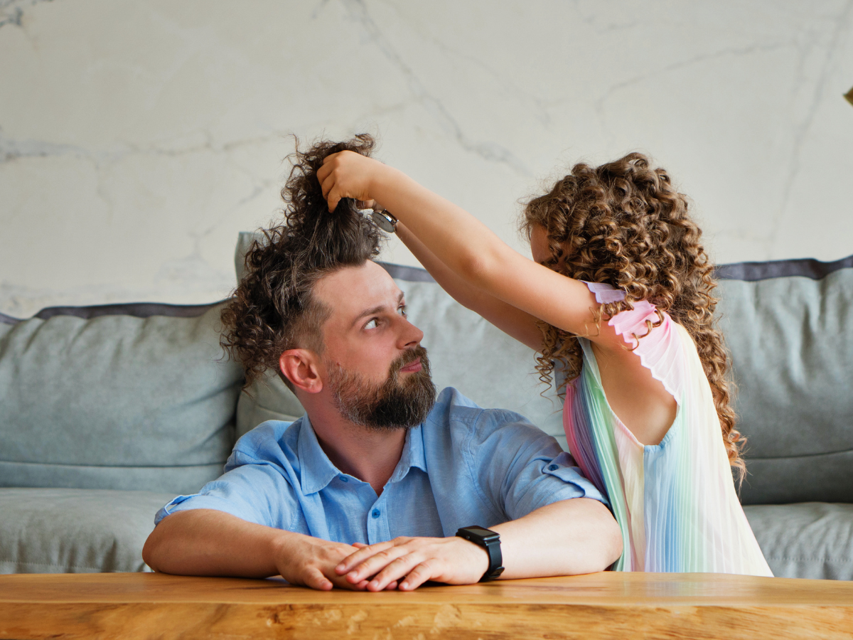 Dad sitting on the floor at coffee table watching his daughter play with his hair, depicting parental burnout and everyday parenting moments