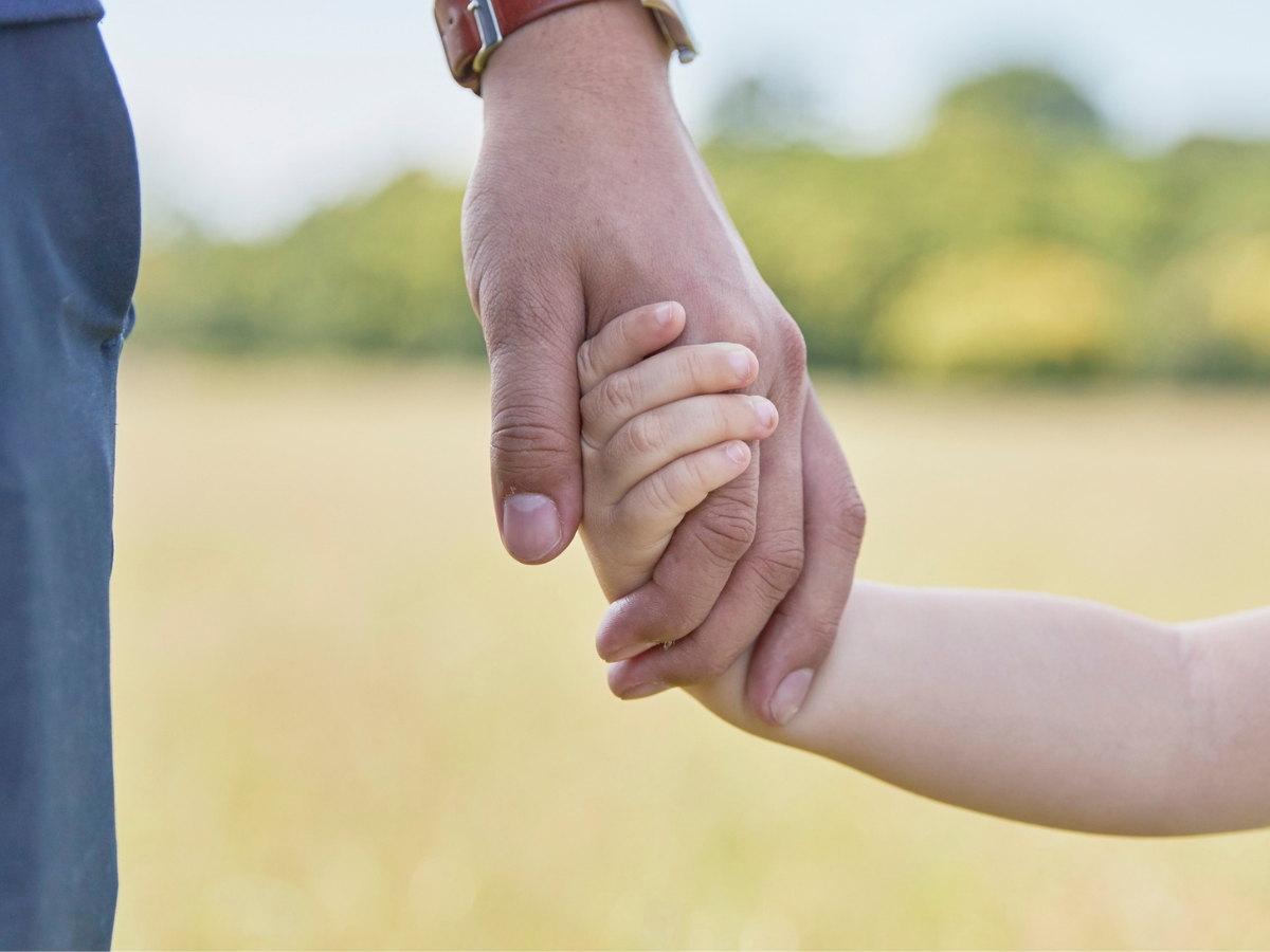 Dad holding his young son's hand with blurred outdoor background, symbolizing parental connection and support