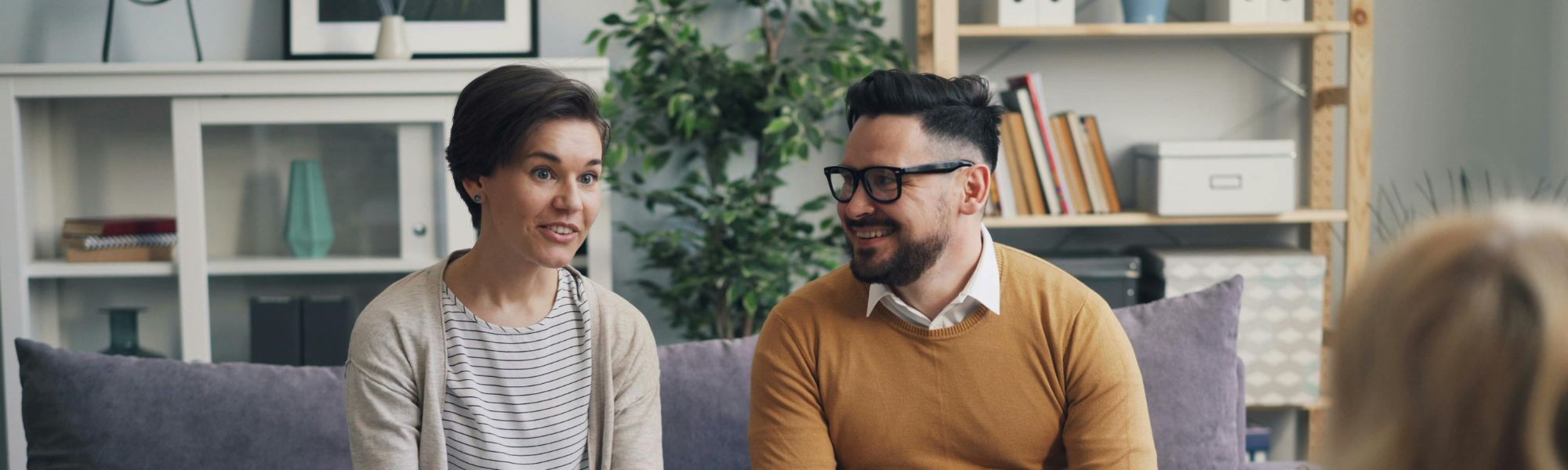 Couple in a therapy session, woman speaking to husband who smiles, therapist partially visible in the background, friendly and supportive environment.