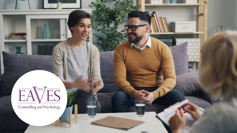 Couple in a therapy session, woman speaking to husband who smiles, therapist partially visible in the background, friendly and supportive environment.