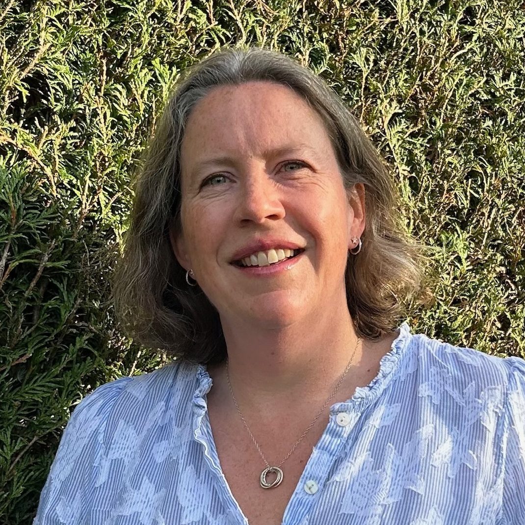 Counsellor in Farnham wearing a blue and white top, standing in front of a green hedge, offering a calm and supportive environment for children, young people, and adults