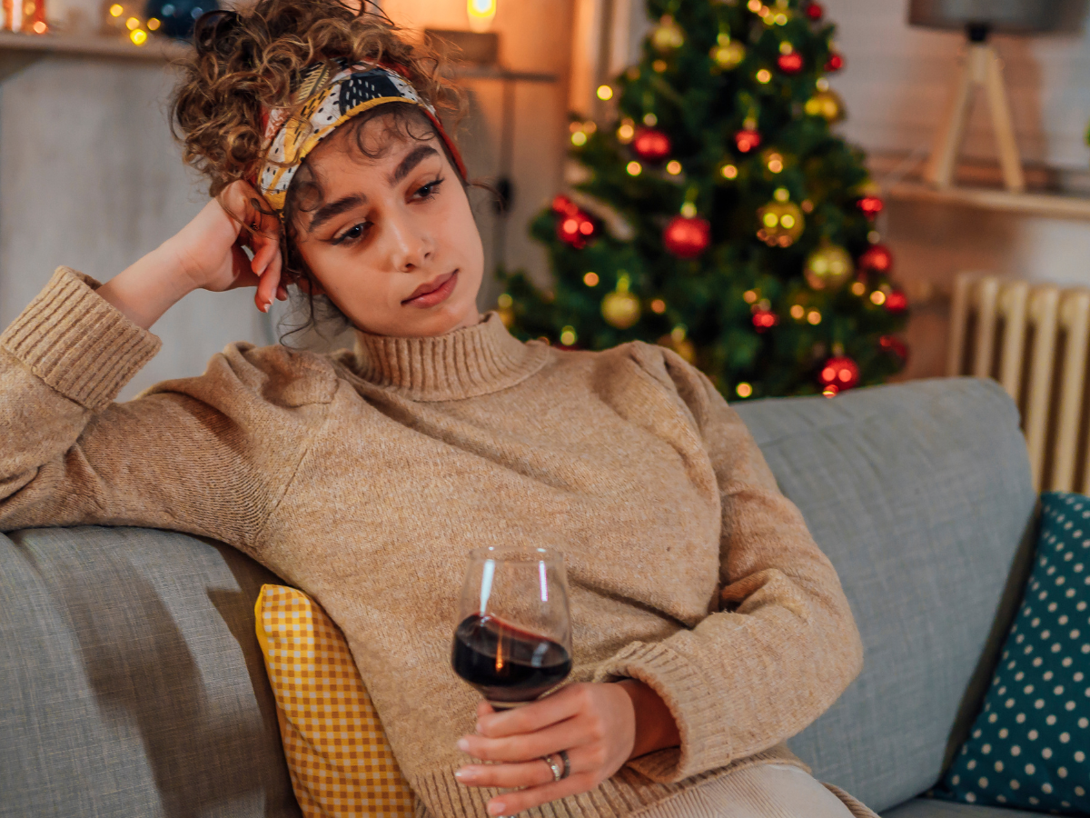 Woman sitting on a sofa with a glass of wine, resting her head on her hand, with a Christmas tree in the background.