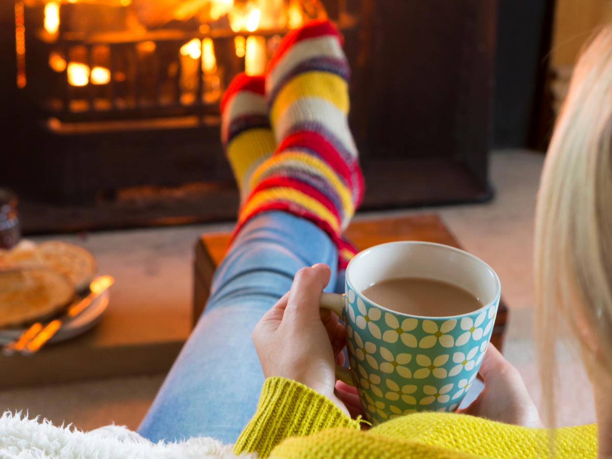 Woman holding a coffee cup while sitting on a sofa in front of a cozy fireplace during the holidays.