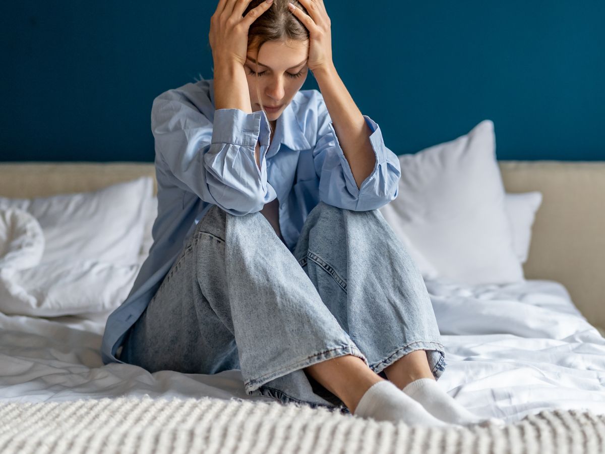 Young woman sitting on a bed, elbows on knees and head in hands, appearing distressed and hopeless.