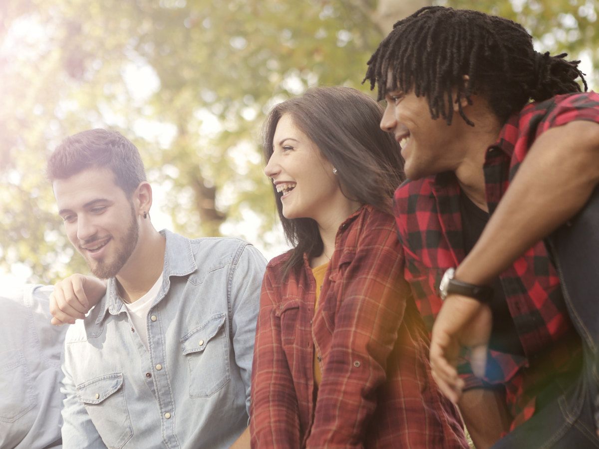 Close-up of three friends—two men and a woman—socializing outdoors, smiling and laughing with blurred trees in the background