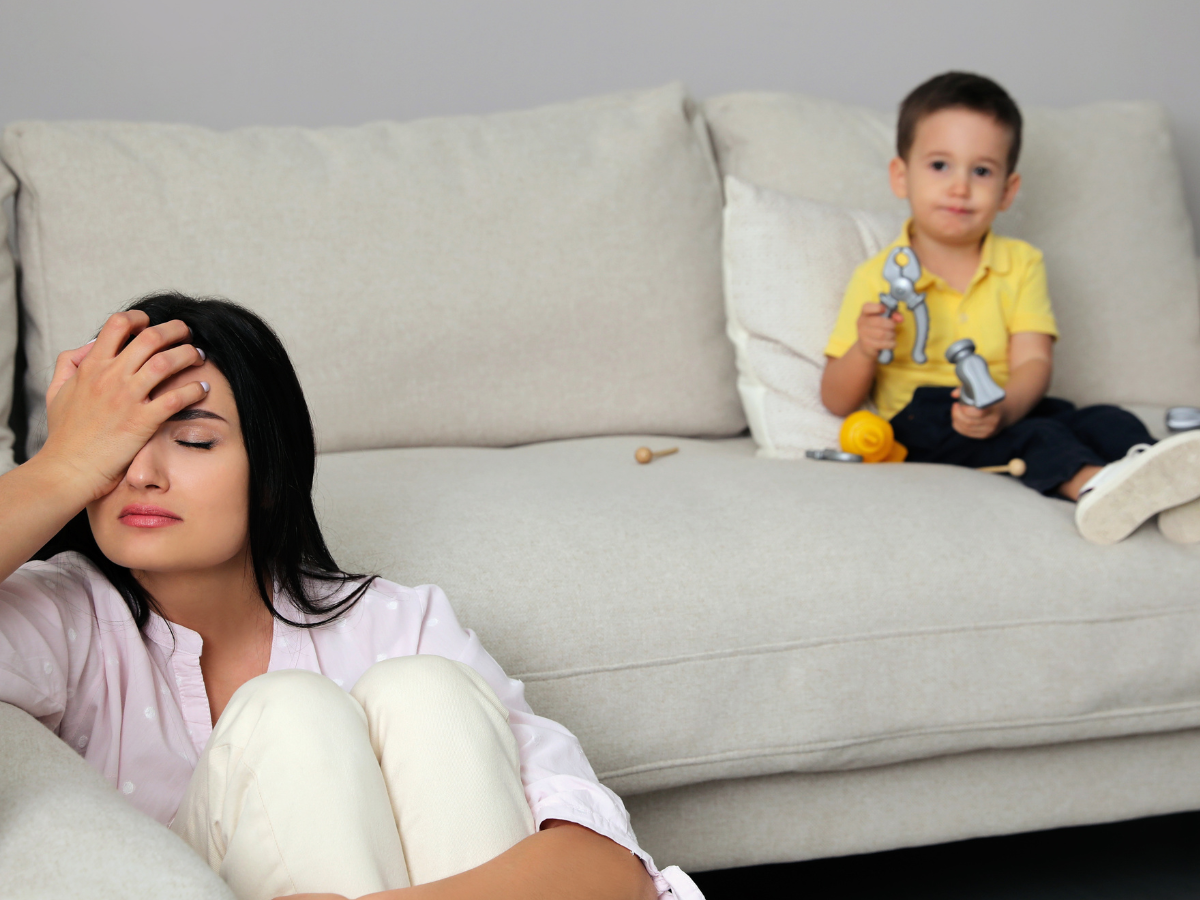 Stressed mother sitting on the floor with head in hand while her child sits quietly on the sofa behind her, symbolizing parental struggles with supporting an anxious child.