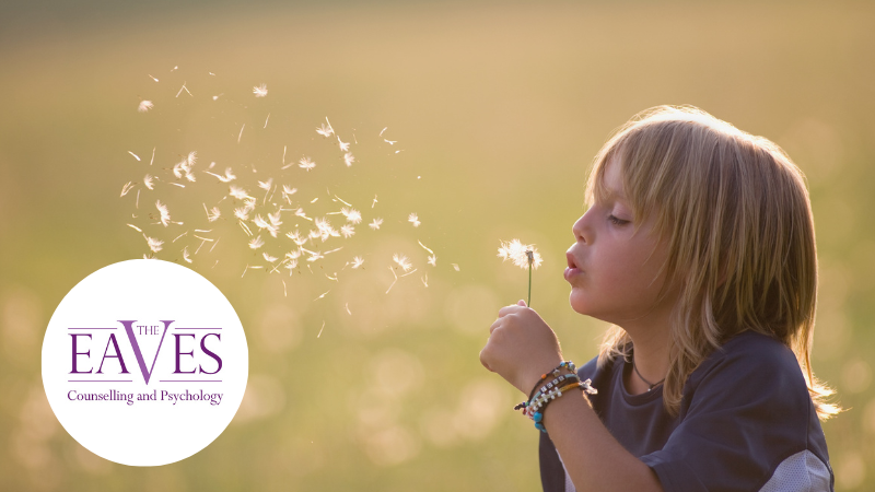 Child blowing dandelion seeds into the wind, symbolising growth and hope – The Eaves Counselling and Psychology with The Eaves logo bottom left in a circle