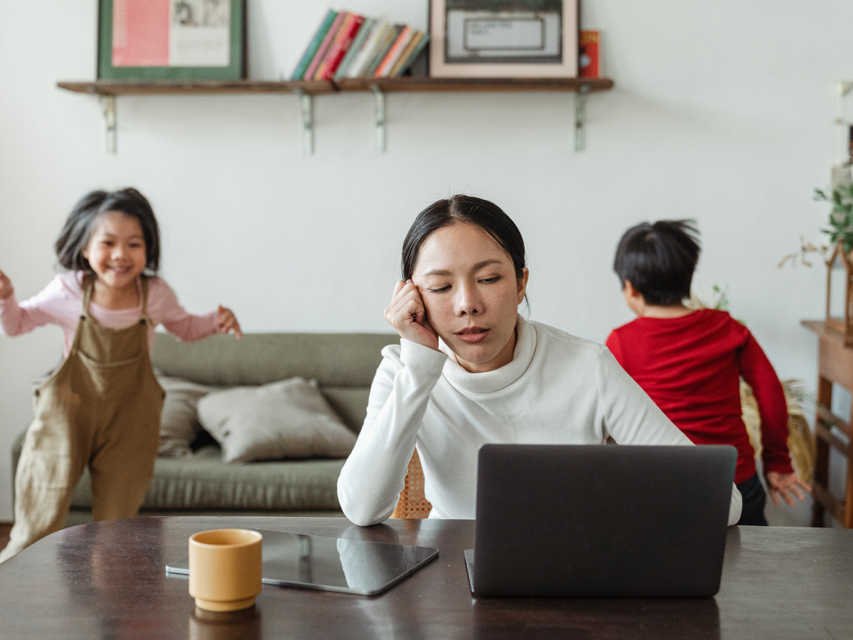 Mother sitting at a dining table with a cup of coffee, resting her head on her hand while looking at a laptop, as children play in the background, illustrating parental stress and emotional exhaustion.