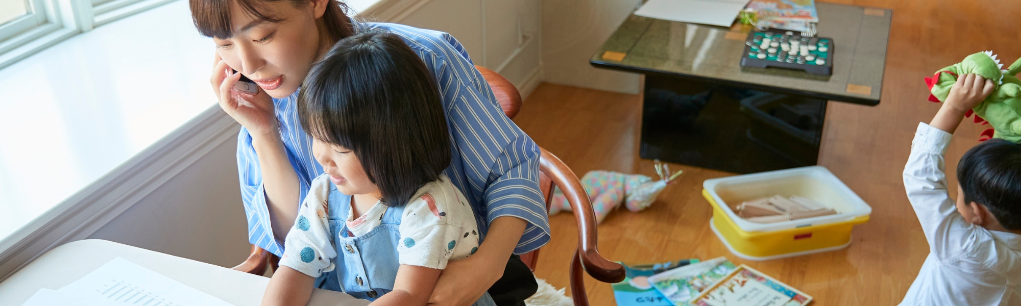 Mom on the phone while holding daughter on her lap, with young son playing on the floor in a cozy living room – illustrating parenting and supporting children with anxiety.