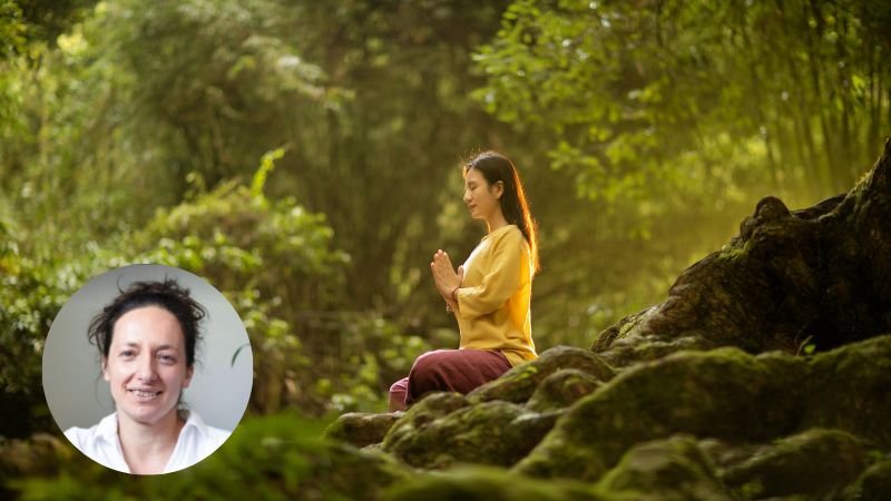 Woman sitting cross-legged in a forest by a river, hands together in a meditation pose