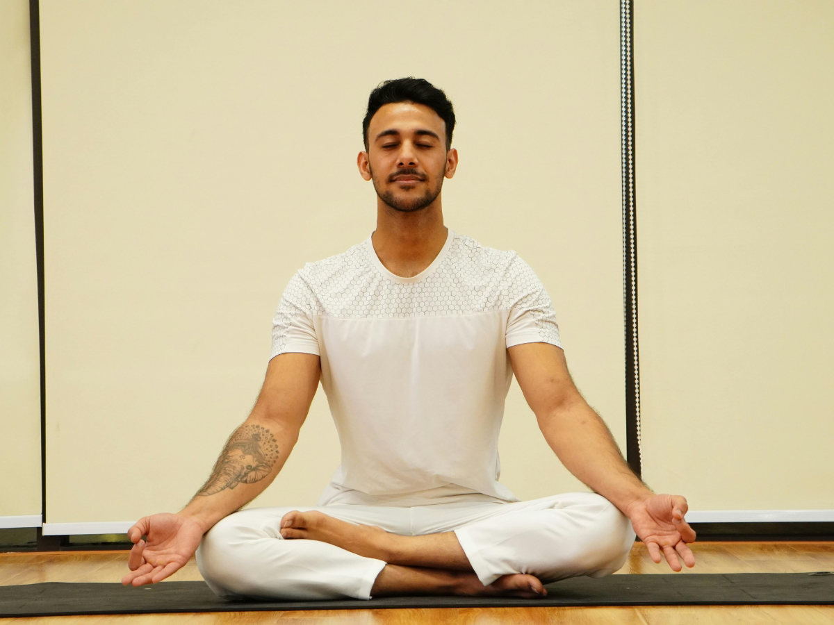 Man sitting cross-legged on a yoga mat in a studio, hands resting on knees in a meditation pose