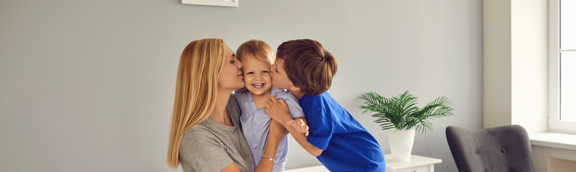 Mom holding toddler while older child kisses toddler’s cheek in playful family moment