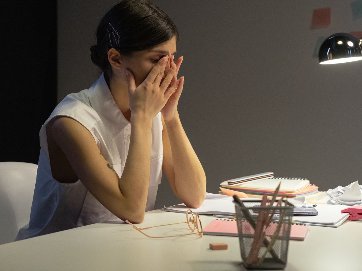 Overwhelmed mom sitting at desk with hands on head surrounded by notes and notepads under desk lamp