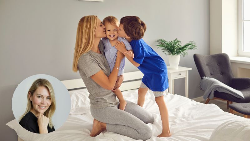 Mom holding toddler while older child kisses toddler’s cheek in playful family moment