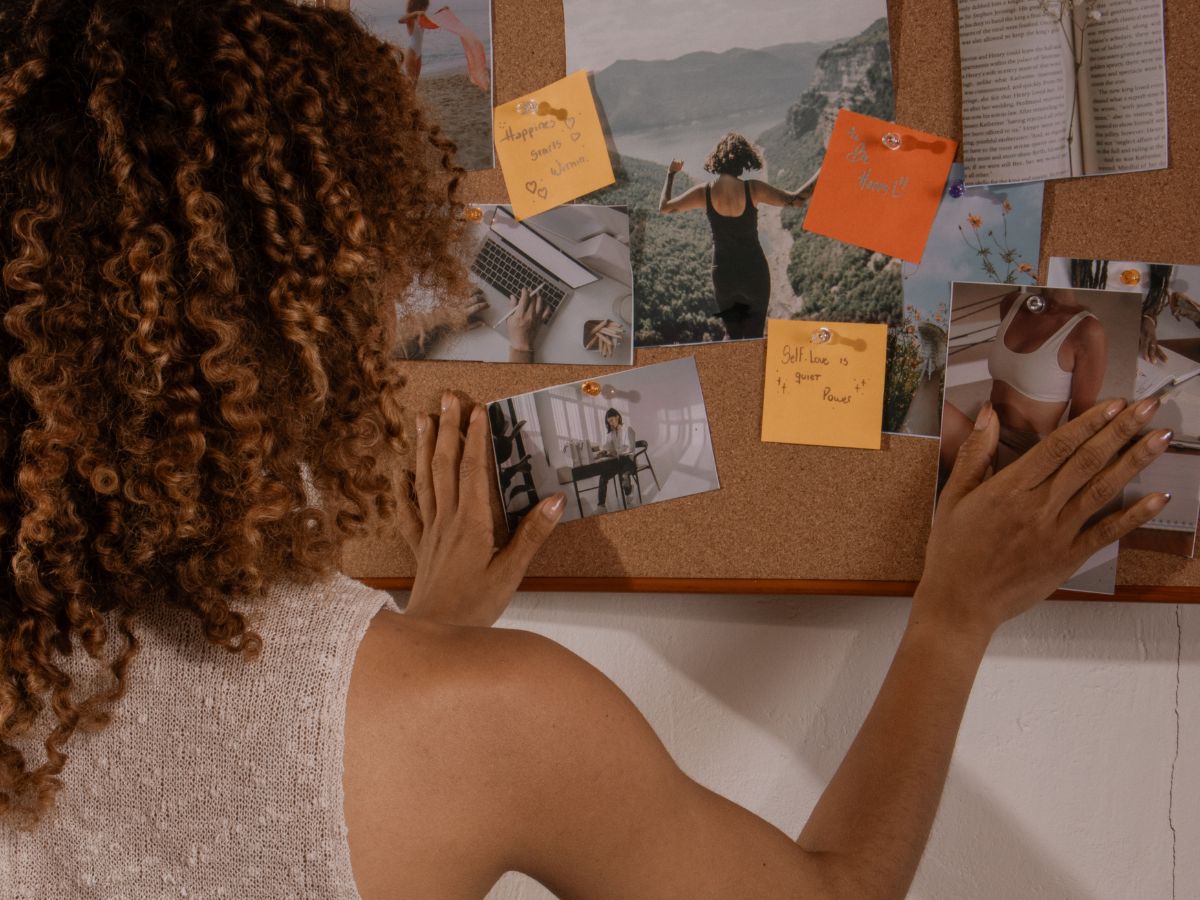 Woman with brown curly hair organising pictures and sticky notes on a pin board, representing reflection and self-discovery.