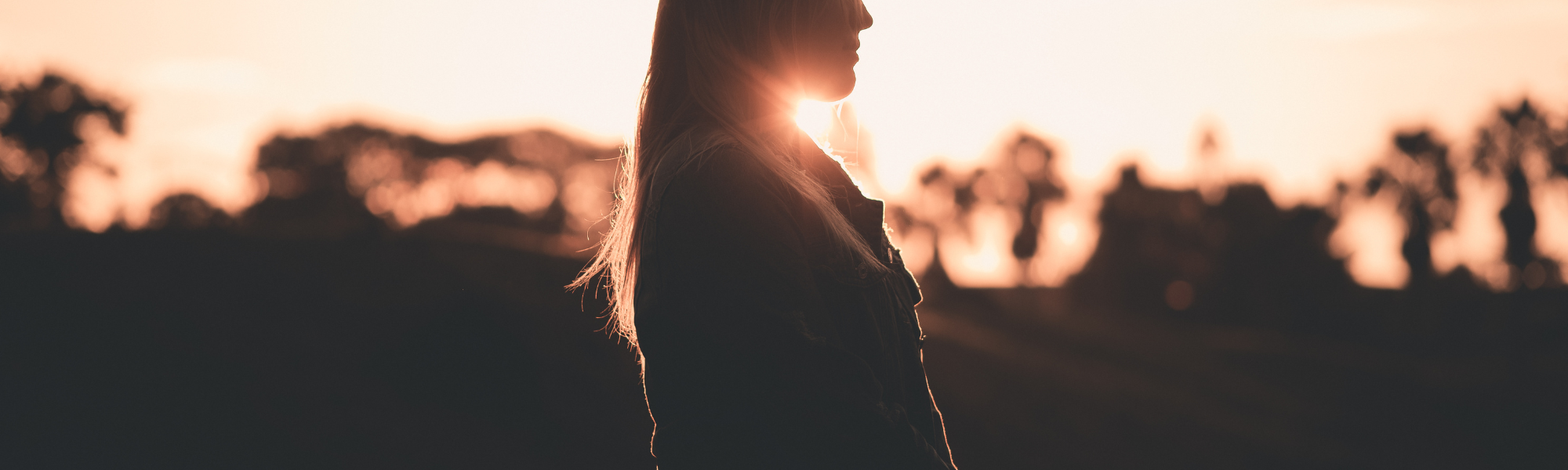 Silhouette of a woman looking away at a glowing sunset over a forested landscape, symbolizing self-reflection and building self-esteem.