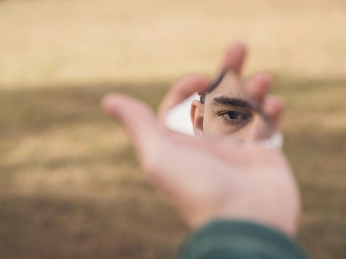 Man holding a broken mirror reflecting his face against a blurred background, symbolising low self-esteem and self-reflection